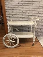 Full view of antique wooden tea cart with distressed white paint and large spoked wheels on hardwood floor in front of a white brick wall.