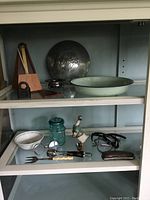 Photo of two shelves with assorted vintage and antique items, including metronome, Fargo hubcap, silver bowl, enamel bowls, blue glass jar, ceramic duck, utensils, and spectacles.
