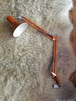Full view of orange adjustable metal desk lamp with screw clamp base, lying on a cream-colored fur rug.