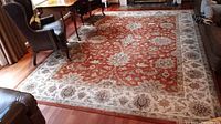 View of the full area rug placed in a living room setting, showing the overall red central area and beige floral border.