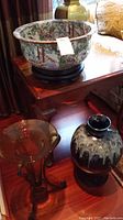 Asian style bowl with detailed floral and scenic patterns on wooden stand and two floor vases below it; one tall amber glass vase with metal stand and one brown ceramic vase