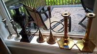 Nine candle holders displayed on a windowsill: two horn style with gold tones and intertwined loops, two gold tone hexagonal base holders, and one black pillar holder.
