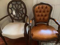 Two chairs side-by-side: one cream upholstered wooden framed chair with geometric open back and one brown vinyl upholstered ornate wooden framed chair.