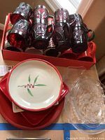 Ten red glass mugs with silver rims and etched star patterns stacked in box, red serving trays beneath ceramic holly bowls and clear glass serving trays next to them