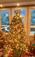 Artificial Christmas tree set up and decorated with warm white string lights in front of window with snow outside. Decorations on tree not included.