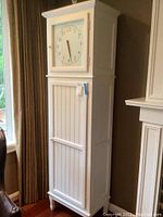 Full view of tall white wooden cabinet styled as a clock tower with clock face door on top and larger lower door below.