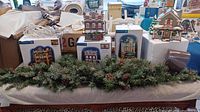 Full view of the set showing multiple Department 56 Snow Village building figurines on their boxes and green faux garland in front