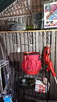 Photo of a Black & Decker orange string trimmer hanging, black metal wire shelving with various items, including coiled wire fencing and packages.