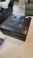 Square brown leather ottoman with button tufted top, showing overall condition and size proportion beside couches.