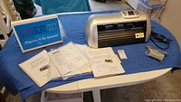 Full lot showing Silver Bullet die cutting machine, manuals, paperwork, power cord, and accessory pouch on a blue cloth surface.