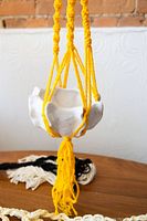 Yellow macrame plant hanger holding white ceramic pot with scalloped edge, displayed hanging.