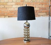 Full view of the lamp with bubble glass base and black shade on a wooden table against a light wall.