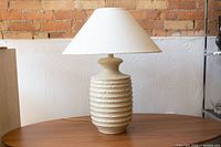 Ceramic table lamp with fabric shade, showing full lamp from side on wooden surface with brick and white textured wall behind.