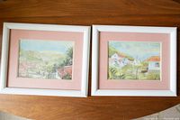 Two framed watercolor artworks with white frames and pink mats, depicting landscapes with buildings and greenery.