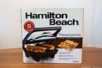View of the front of the box showing Hamilton Beach branding, model number 25460C, image of the panini press with sandwiches inside, and a 5-year warranty badge.
