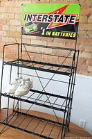 Black metal three-tier display rack with Interstate Batteries metal sign attached at top. Shoes placed on second tier for scale.