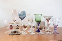 Lot of 11 assorted vintage stemmed glasses including colored, clear, and decorated types, shown on wooden surface against white wall.