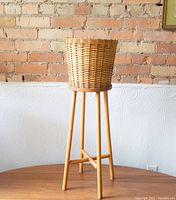 Full view of the wicker planter basket on a tall wooden stand against a white embossed wall and exposed brick background.