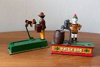Photo showing two metal dog trick coin banks side by side on a wood surface with a white wall background.
