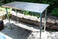 Front angle view of stainless steel storage table with two wire shelves, outdoors on a patio.