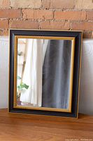 Full front view of vintage mirror showing black frame and gold inner detailing against brick wall background.