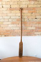 Full view of the vintage wooden paddle leaning against a brick wall and white textured surface.