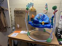 Full view of the Baby Einstein Neptune's Ocean Discovery Jumper showing the seat, attached ocean-themed interactive toys, and original box on the table in a room with brick walls.