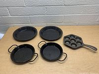 Set of one cast iron muffin pan with seven molds and four black enamel covered camping plates (two round, two handled skillet-style).