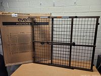 Photo of dark wood framed metal mesh baby and pet gate extended, leaning against wall, with original Evenflo box behind.