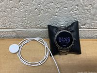 Overall view of the Fossil smart watch displayed on a black pillow beside a white magnetic charging cable, showing the screen active with time displayed.