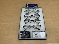 Five pairs of Gamma Ray Optics reading glasses displayed on a white foam tray against a wooden surface. Various frame styles and colors visible; glasses are marked +3.50 strength. Tray is used for display only and is not included.