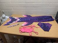 Photo showing purple and pink mermaid tail fabric, purple swim bottoms, pink bikini top, and floral headband laid flat on table