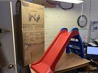 Child's red and blue Little Tikes slide shown standing upright next to its original cardboard box in an indoor room with various office items in the background.
