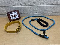 Yellow leather dog collar, blue rope dog leash with padded handle, and Logical Leather product box on a wooden table against a brick wall background.