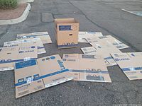 Ten large cardboard moving boxes flattened and one box assembled to show size and condition in outdoor parking lot.