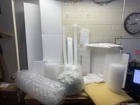 Wide photo showing various sizes of foam sheets standing upright at the back, some foam rolls, and on the table bubble wrap rolls and bags of packing peanuts.