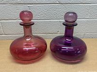 Two glass decanters side by side, one pink and one purple, showing the overall shape and glass color.