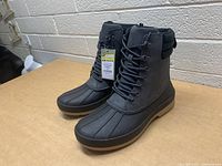 Side angle view showing the pair of black Goodfellow boots with tags attached, size 10, on a table against a wall.