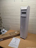 White bathroom tissue storage cabinet and dispenser displayed standing on a table against a beige brick wall, with instruction manual visible.