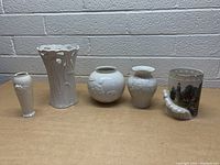 Five Lenox porcelain vases and one painted glass decorative item arranged on a table against a cinderblock wall.