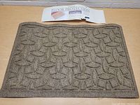 Top-down photo of rectangular brown Weather Guard entry mat with interlocking oval geometric pattern, label visible showing floor protection purpose.