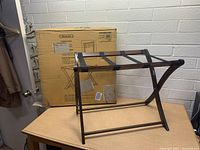 Photo of new Scarlett luggage rack with wooden frame and black fabric straps, placed on a table with original box in background.