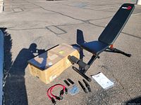 Pelpo adjustable workout bench partially assembled with displayed accessories and instruction manual beside a cardboard box outdoors in sunlight