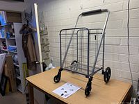 Full view of folded shopping cart on a table in a room, showing metal frame, handle, wheels, and basket.