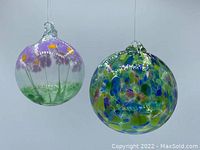 Two handmade glass balls with hanging loops. One ball features purple flowers with yellow centers and green stems in a translucent design. The other exhibits a blue, green, purple, and white stained glass effect pattern.