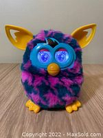 Front view of Hasbro Furby Boom showing magenta and teal green houndstooth faux fur, bright yellow ears, feet, beak, and lighted blue eyes.