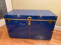 Front closed view of large blue metal steamer trunk with brass accents placed on floor near door.