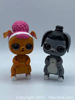 Front view of two LOL Surprise interactive pet figures standing upright. Left figure is orange with pink hair and pink hat, right figure is gray with dark bun hair. Both have large eyes and detailed facial features.