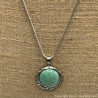 Front view of the silver tone chain necklace with the round green jade pendant on a textured brown background.