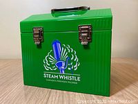 Front angle of green Steam Whistle metal box showing logo, silver handle and latches